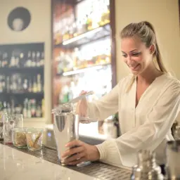 Uśmiechnięta barmanka przygotowuje drinka, ucząc się, jak zostać barmanem. Za nią półki z alkoholem.