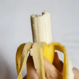 Dłoń trzyma obranego banana, który jest częściowo zjedzony. Czy banany są kaloryczne? Ten owoc może być smaczną przekąską.