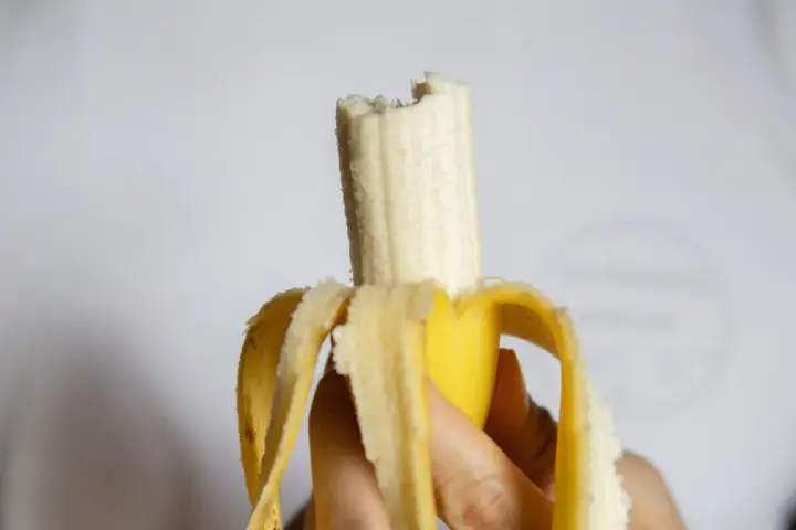 Dłoń trzyma obranego banana, który jest częściowo zjedzony. Czy banany są kaloryczne? Ten owoc może być smaczną przekąską.